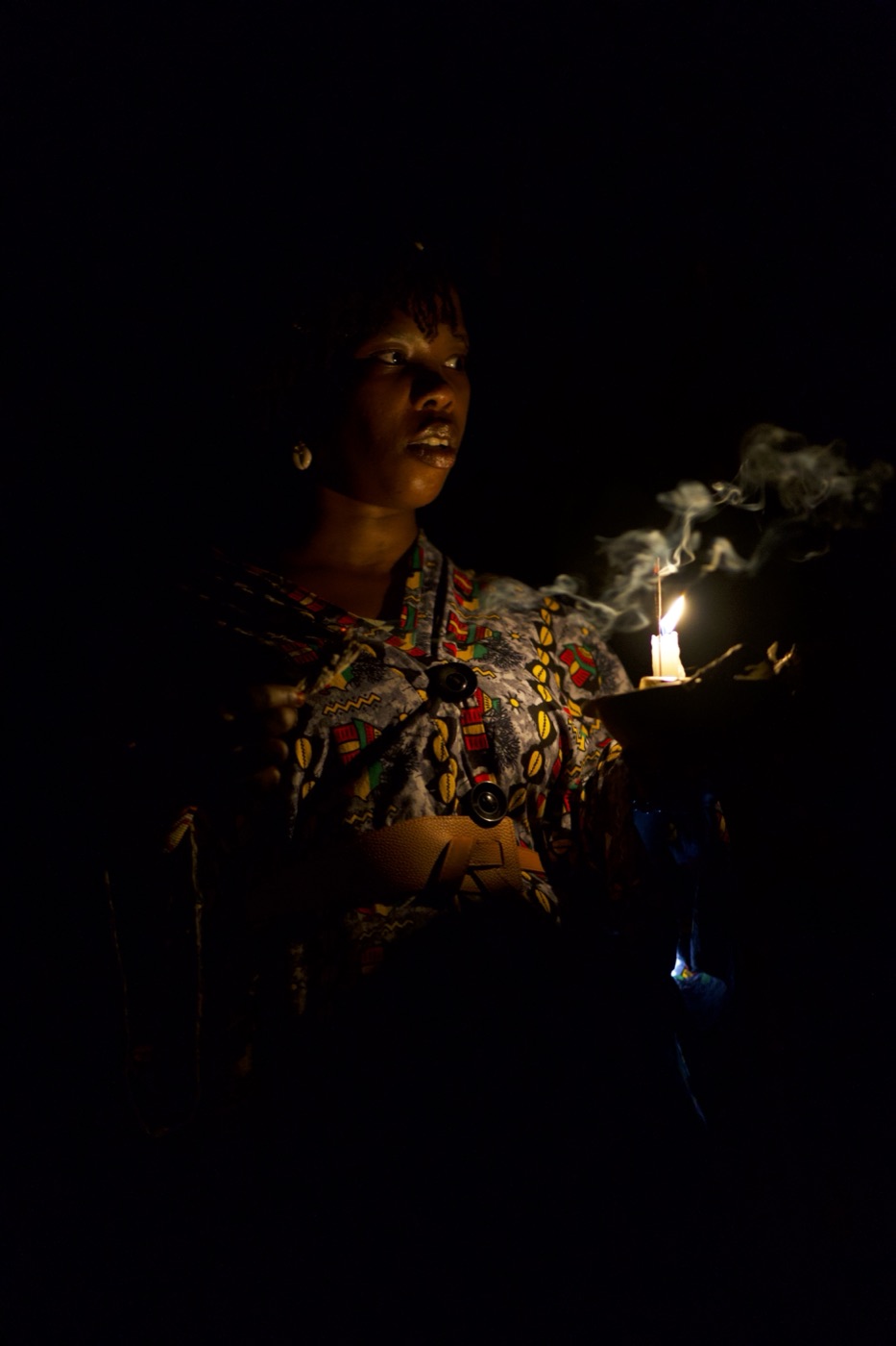 A woman holds a lit candle and incense, smoke rising against darkness.